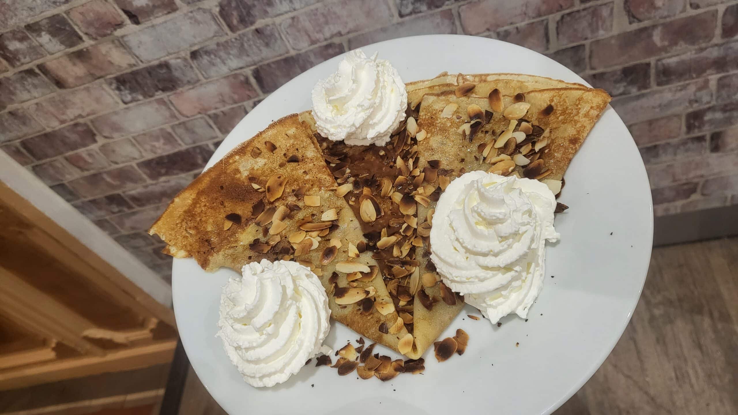 Quatre crêpes dorées aux amandes effilées et trois rosettes de crème chantilly sur une assiette blanche, vue de dessus.