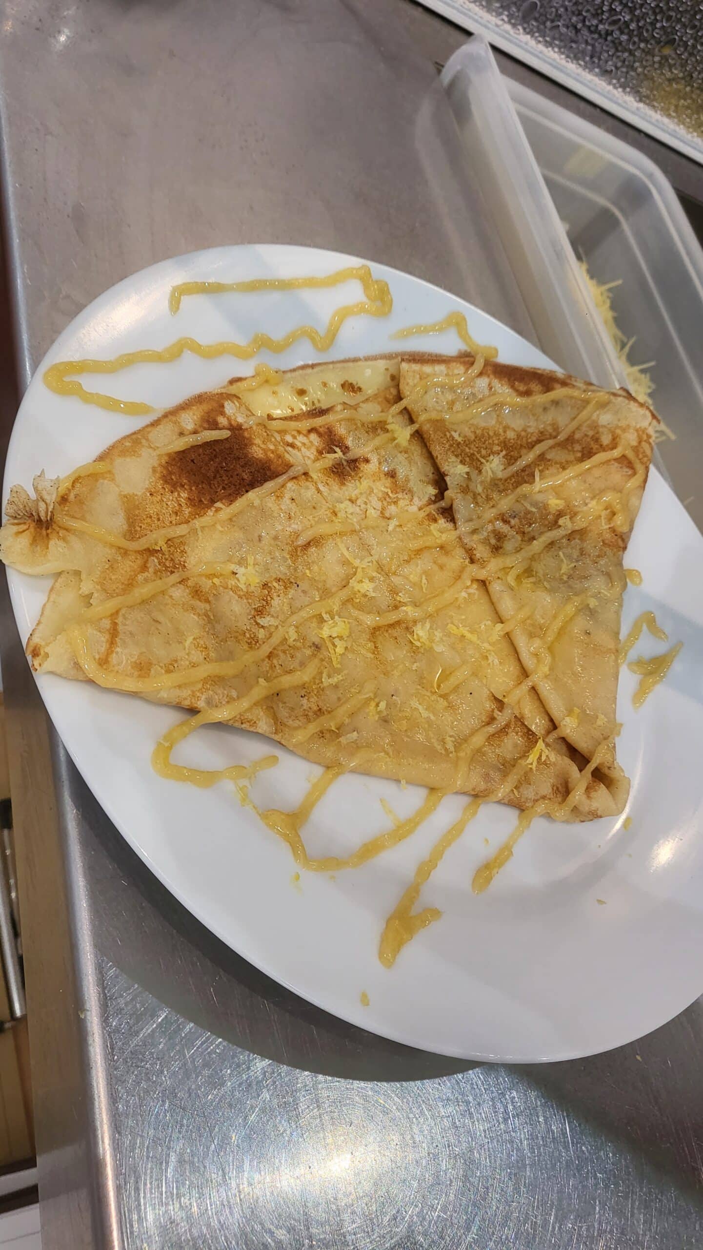Crêpe pliée dorée sur une assiette blanche, nappée d'une sauce jaune et de zeste de citron, posée sur un comptoir en inox.
