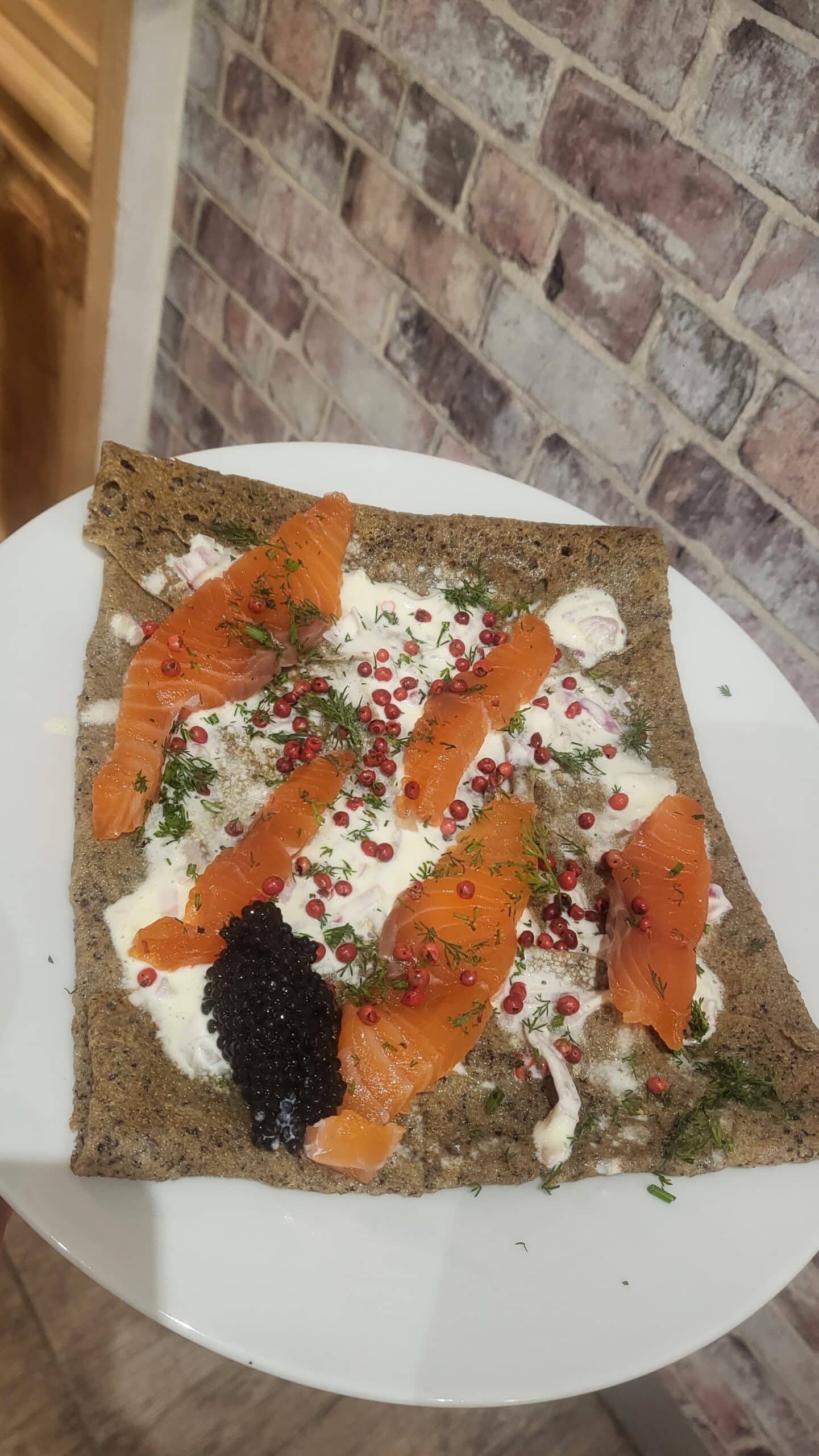Crêpe de sarrasin gourmande garnie de tranches de saumon fumé, caviar noir, crème fraîche, baies roses et aneth, sur une assiette blanche.
