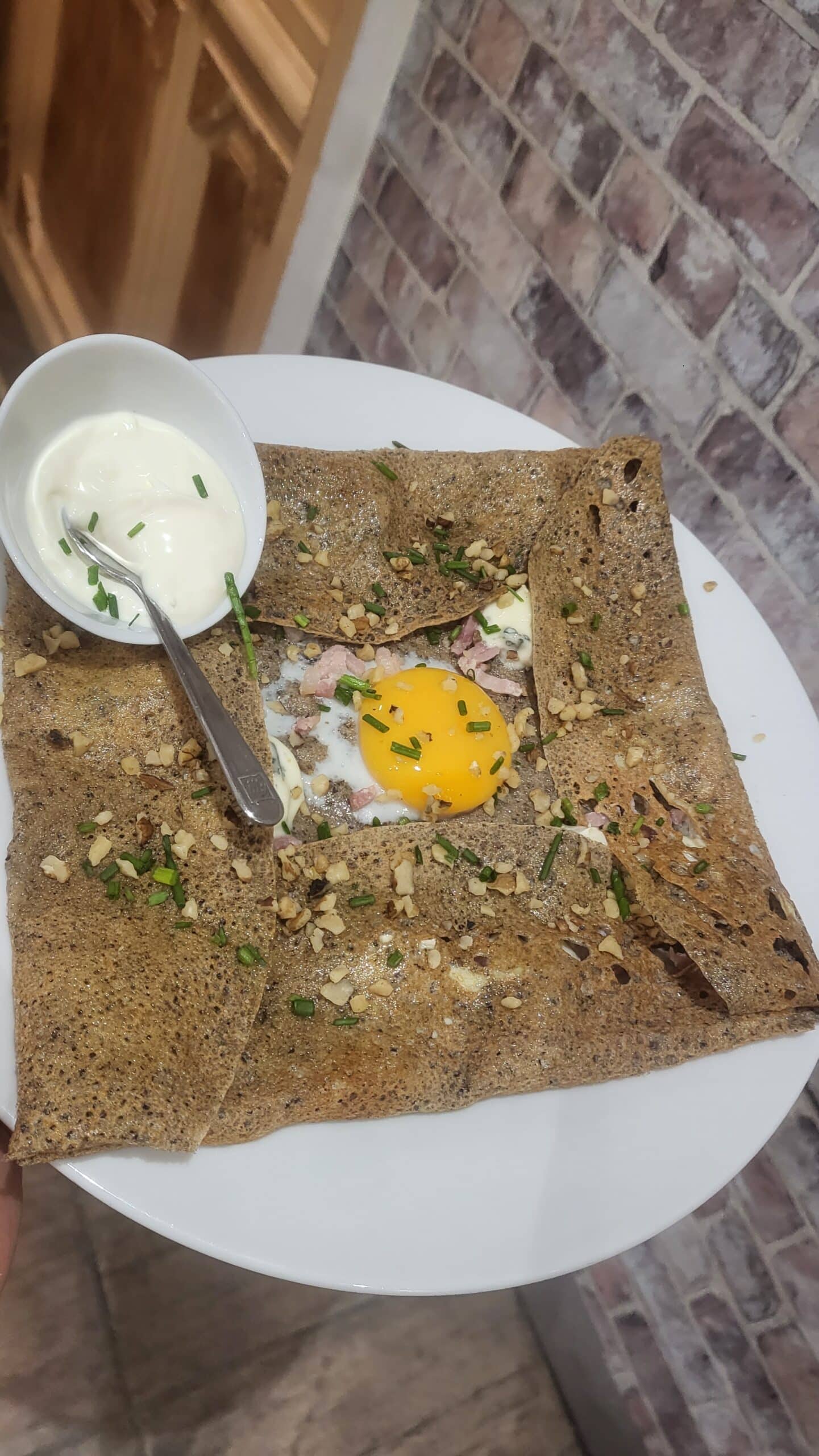 Galette bretonne salée garnie d'un œuf, lardons, fromage, noix et ciboulette, avec crème fraîche en accompagnement.