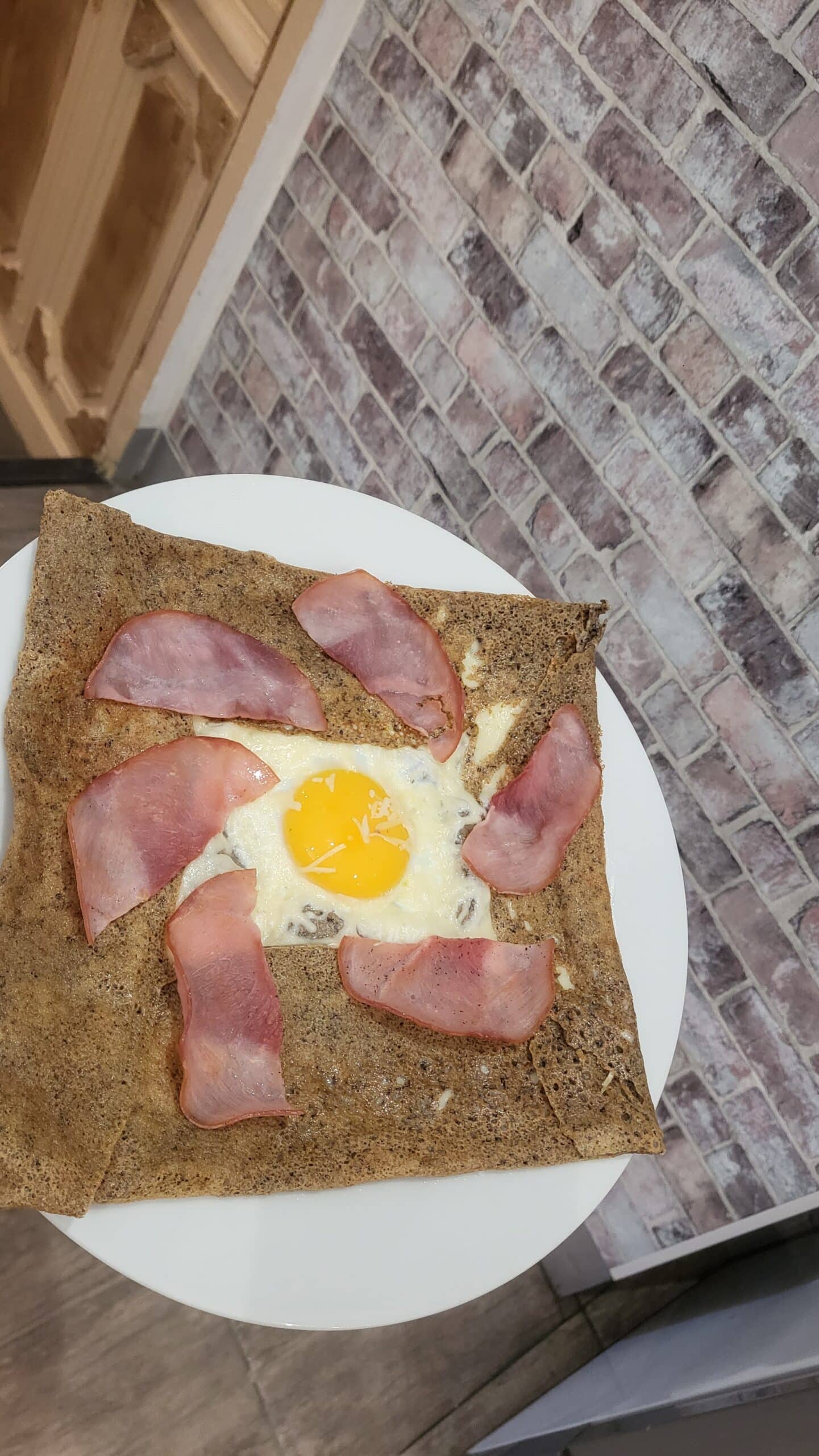 Galette de sarrasin carrée garnie d'un œuf au plat, fromage fondu et bacon. Plat breton servi sur assiette blanche devant mur en briques.