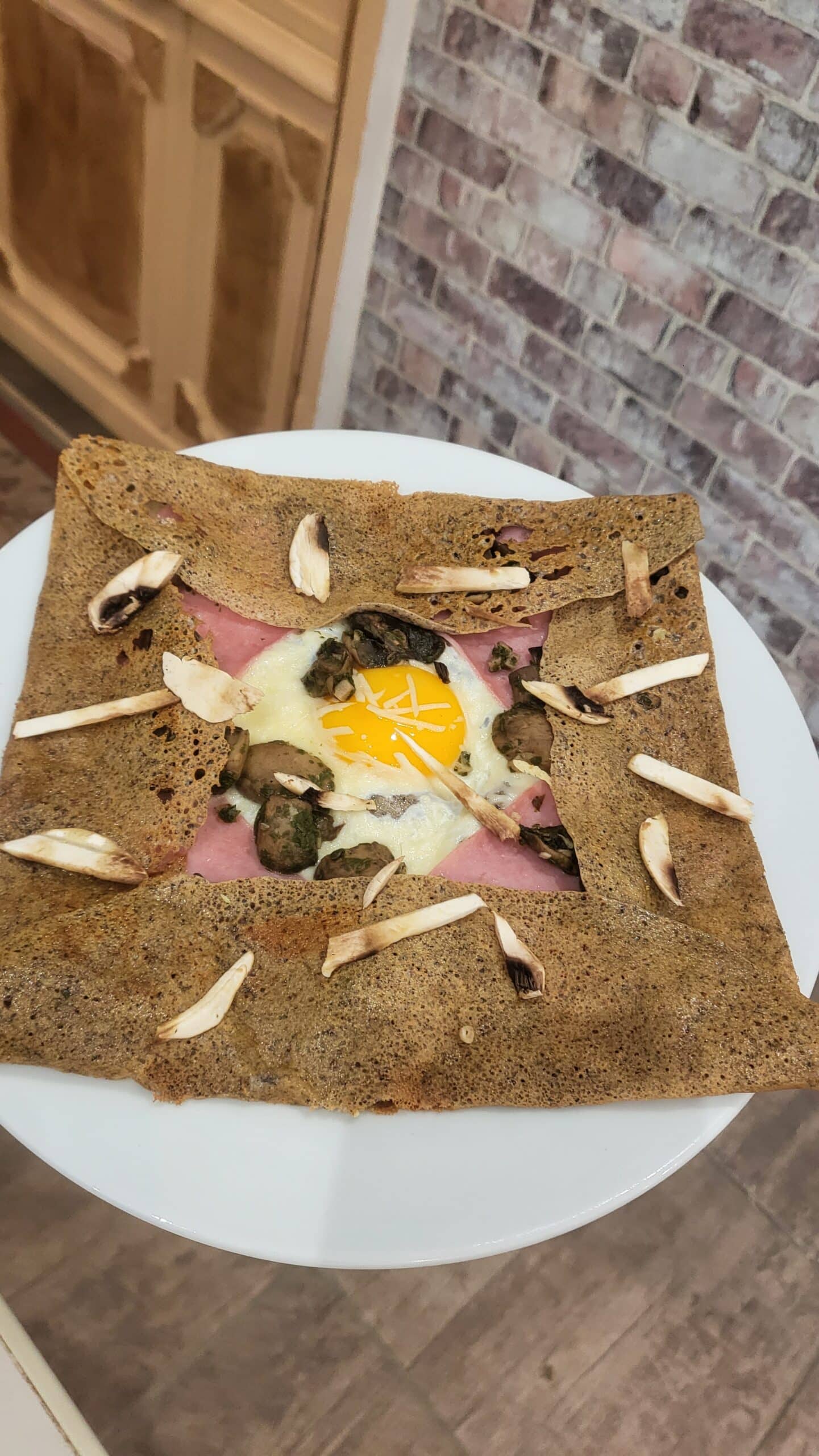 Galette bretonne aux bords repliés, garnie d'un œuf au plat, jambon rose et champignons sautés. Des lamelles de champignons décorent le dessus.
