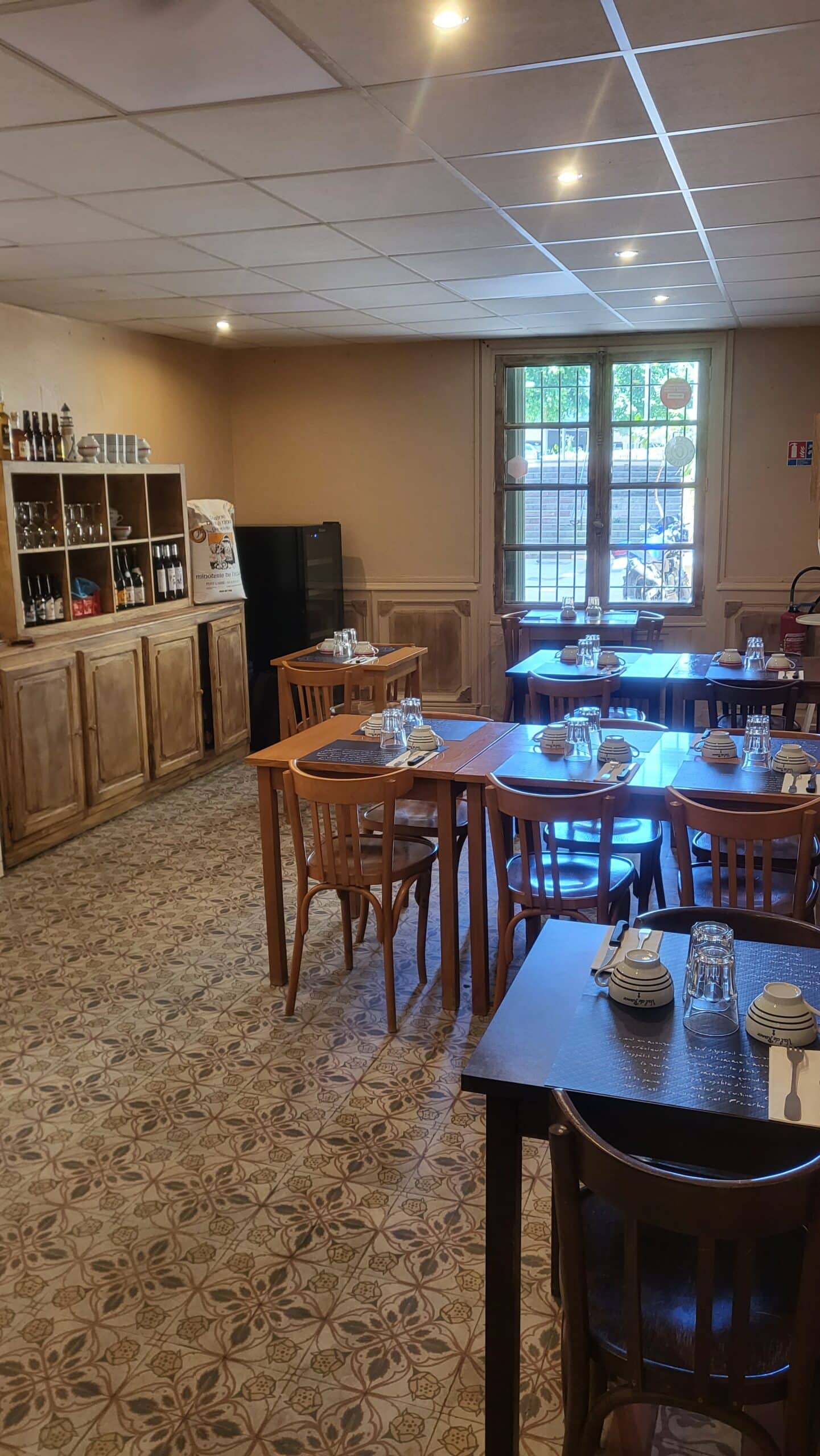 Intérieur de restaurant chaleureux : tables bois dressées Val de Rance, sol carrelé fleuri, armoire Minoterie de Leca, fenêtre. Plafond éclairé.