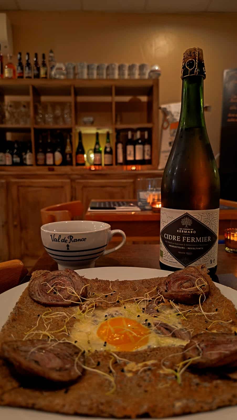 Galette bretonne à l'œuf, viande et germes, cidre fermier Kermabo et tasse Val de Rance en restaurant.
