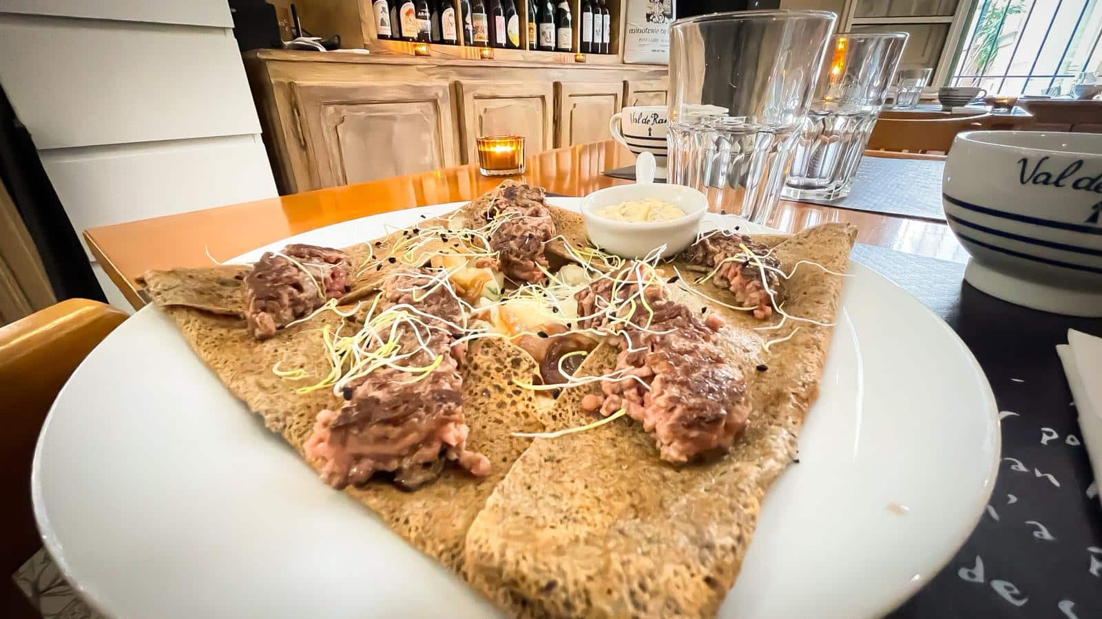 Galette de sarrasin garnie de tartare de viande, œuf et jeunes pousses, sur table de restaurant avec tasse "Val de Ran" et verres.