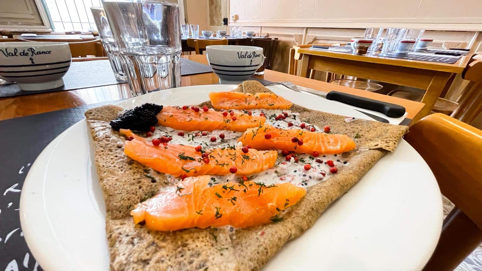 Galette bretonne au saumon fumé et caviar, garnie de baies roses et aneth, sur une table de restaurant avec bol "Val de Rance".
