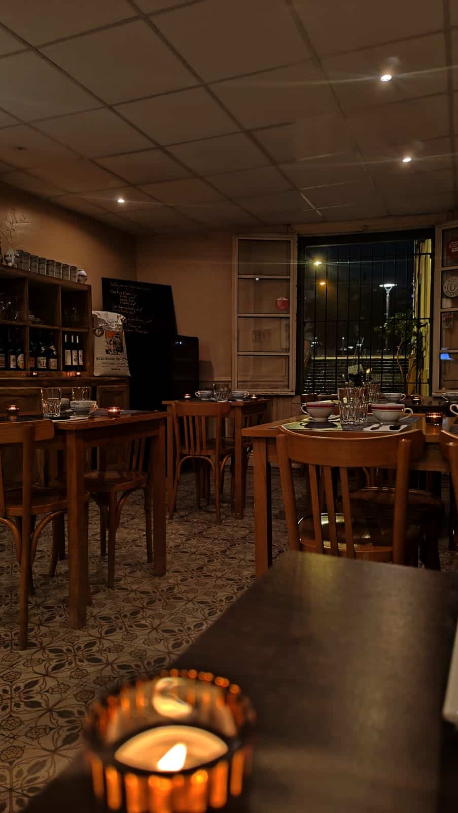 Intérieur de restaurant chaleureux éclairé aux chandelles. Tables en bois, sol carrelé, tableau menu, et vue nocturne sur la rue.