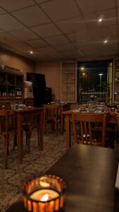 Intérieur de restaurant cosy et tamisé le soir. Tables en bois dressées avec bougies allumées, sol carrelé, et grande fenêtre donnant sur une rue illuminée.