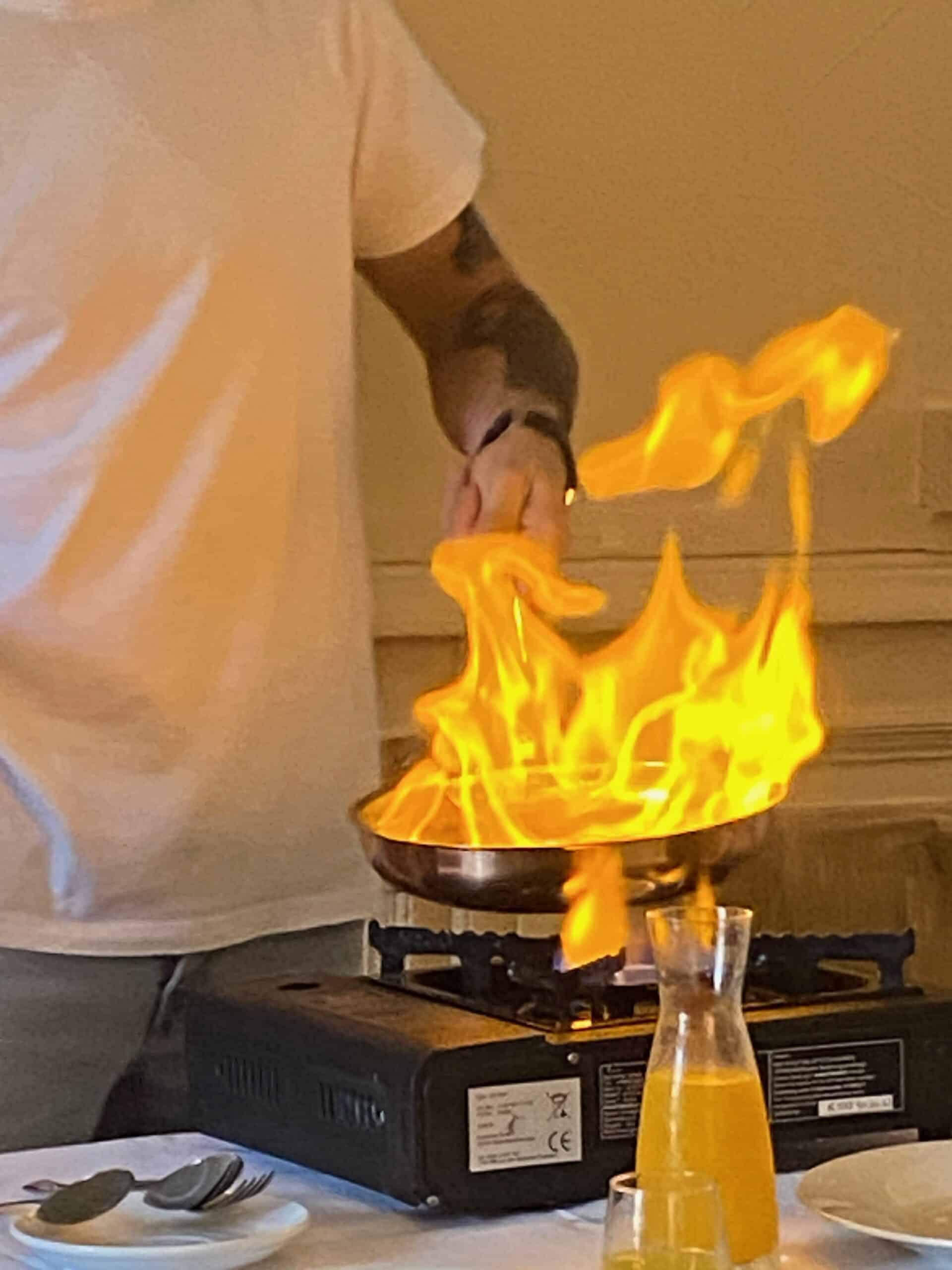 Chef flambant un plat dans une poêle sur un réchaud à gaz portable (CE). Flammes vives illuminent la scène. Table avec assiette, couverts et carafe de jus orange.
