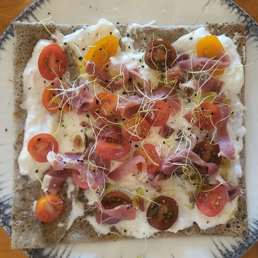 Galette de sarrasin gourmande garnie de burrata crémeuse, tomates cerises multicolores, jambon cru, germes et poivre noir.