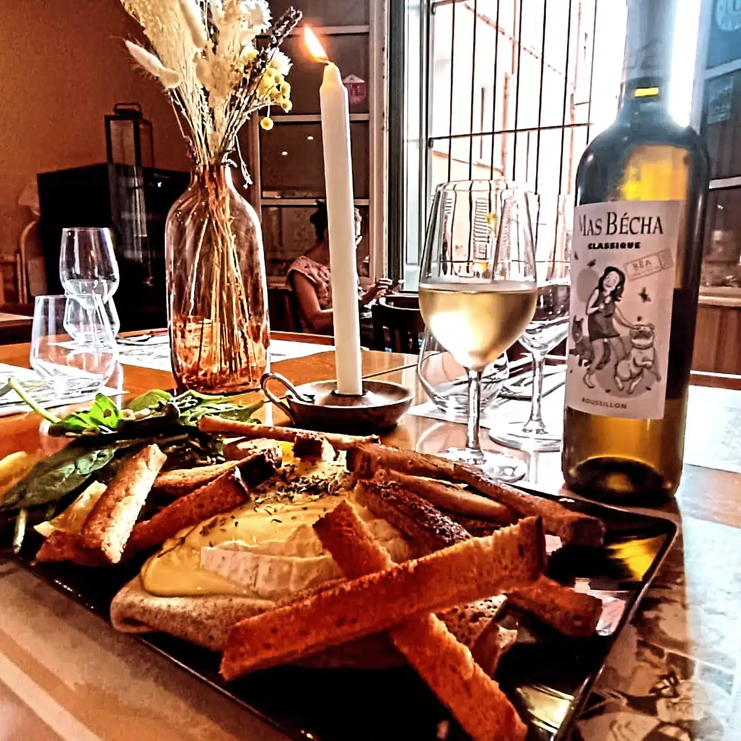 Table de restaurant chaleureuse avec camembert rôti, gressins, salade fraîche, un verre de vin blanc Mas Bécha, et une bougie allumée.