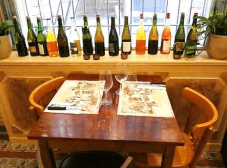 Table de bistrot pour deux, dressée avec verres à vin. En arrière-plan, une rangée de quatorze bouteilles de vin nature ou de cidre colorées.