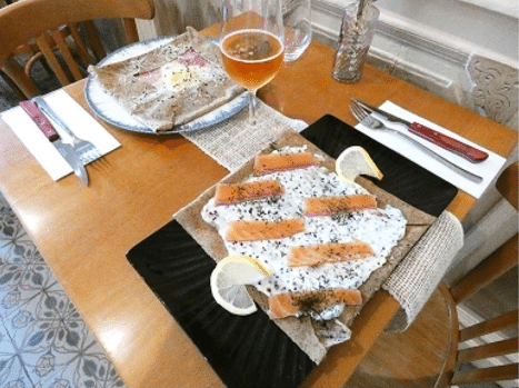 Table de crêperie : galette de sarrasin ouverte au saumon fumé et fromage frais, et galette complète œuf-jambon, avec un verre de cidre.