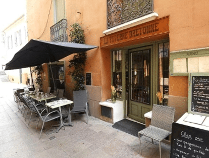 Façade orange de LA TAVERNE BRETONNE, porte verte, avec une terrasse extérieure sous un grand parasol noir dans une rue pavée.