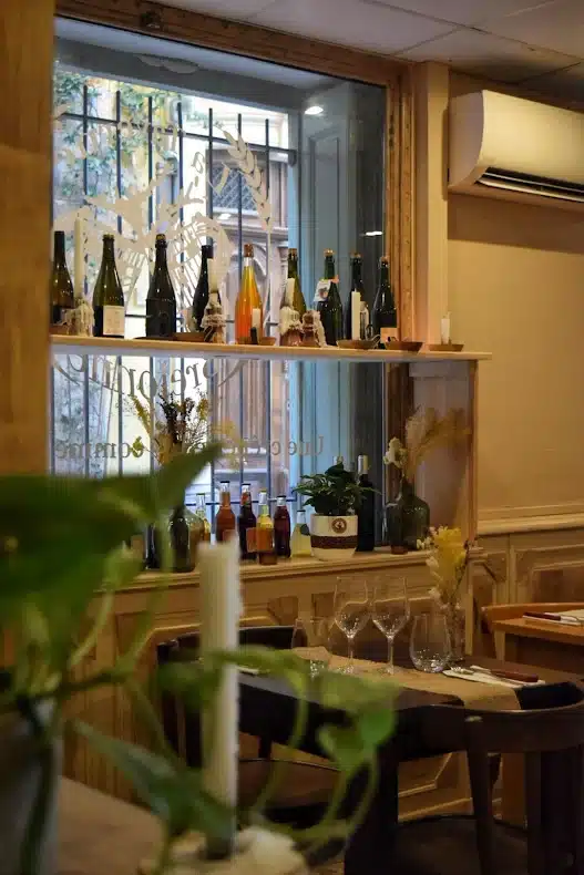 Ambiance de restaurant avec bouteilles de vin, verres sur table dressée et décor végétal devant une fenêtre.