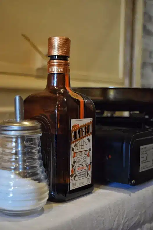 Bouteille de liqueur Cointreau à côté d'un sucrier en verre et d'un réchaud de cuisine portable.