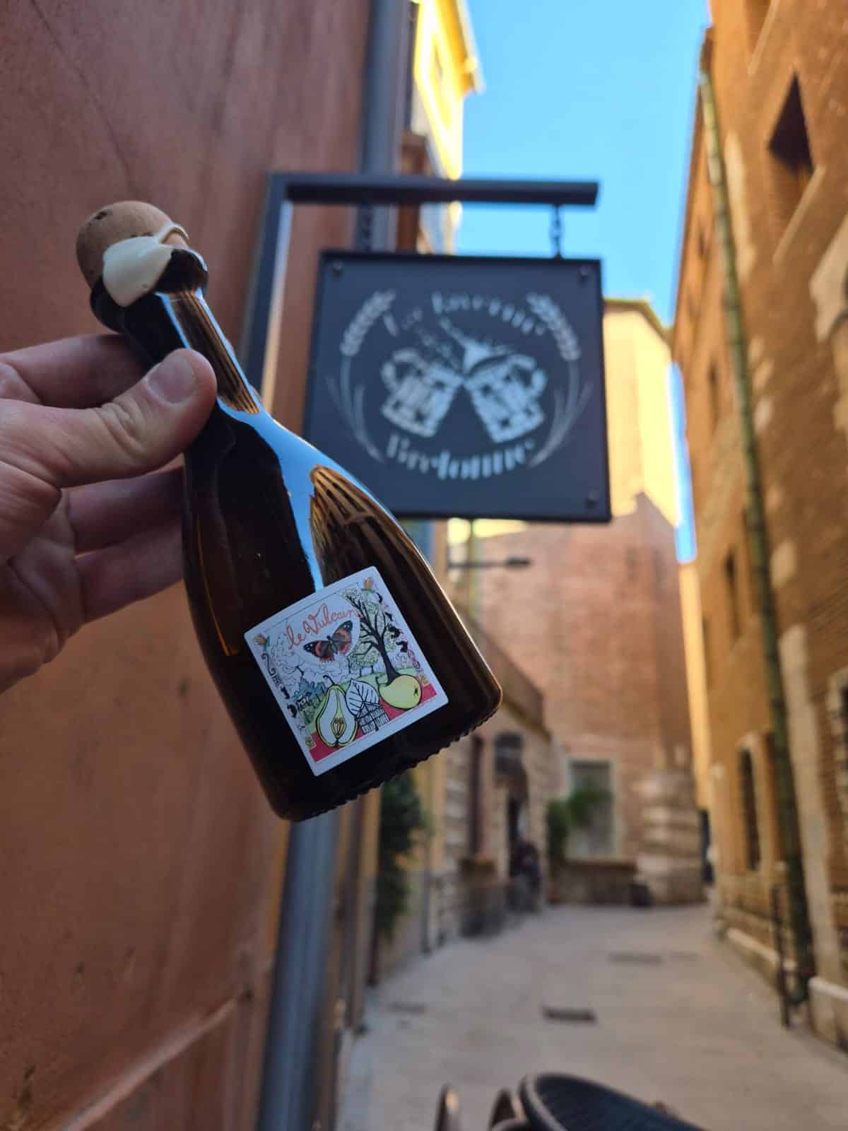 Main tenant une bouteille de cidre Le Vulcain devant l'enseigne d'un bar breton dans une ruelle pittoresque.