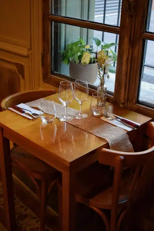 Table en bois pour deux avec verres et chemin de table en toile de jute devant une fenêtre de restaurant chaleureuse.